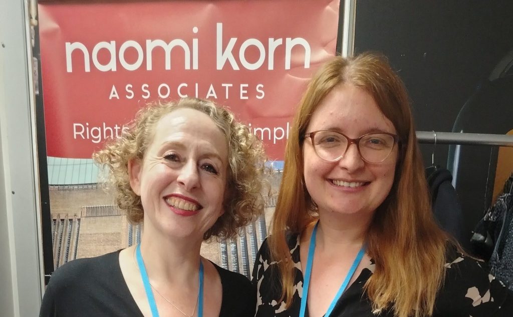Naomi and Jess at the conference, in front of a Naomi Korn Associates banner.